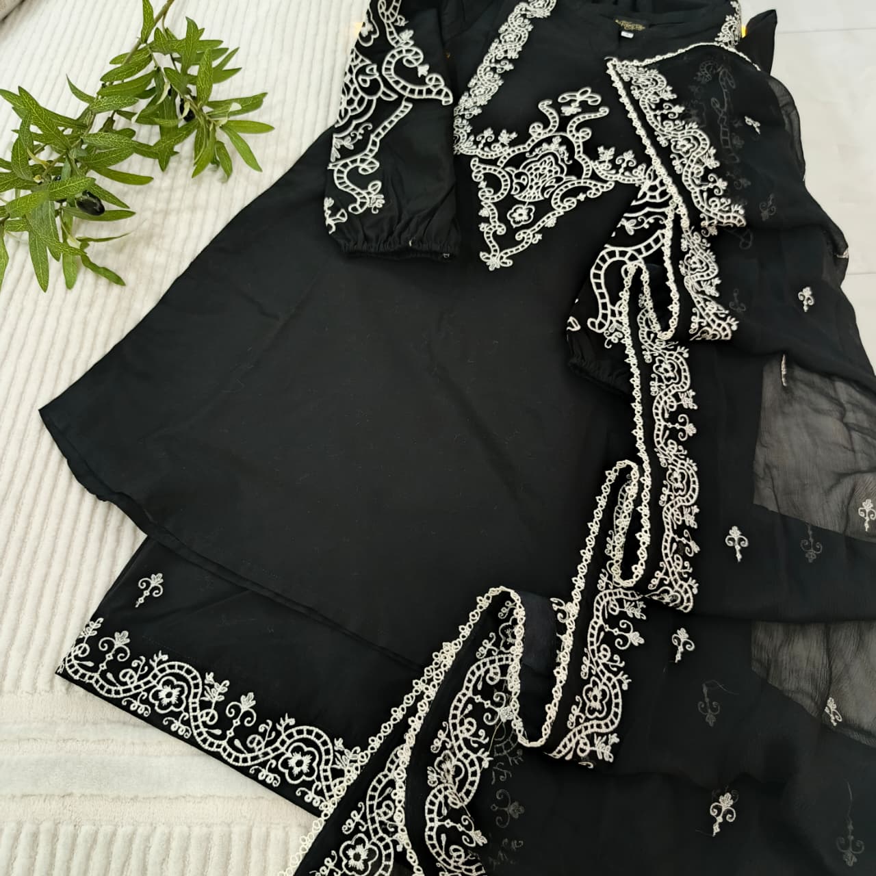 Black garment with white embroidery on a textured surface with green leaves.
Celebrate Eid with Noor-e-Zar Collection - Flat 25% Off Festive Pakistani Dresses Online