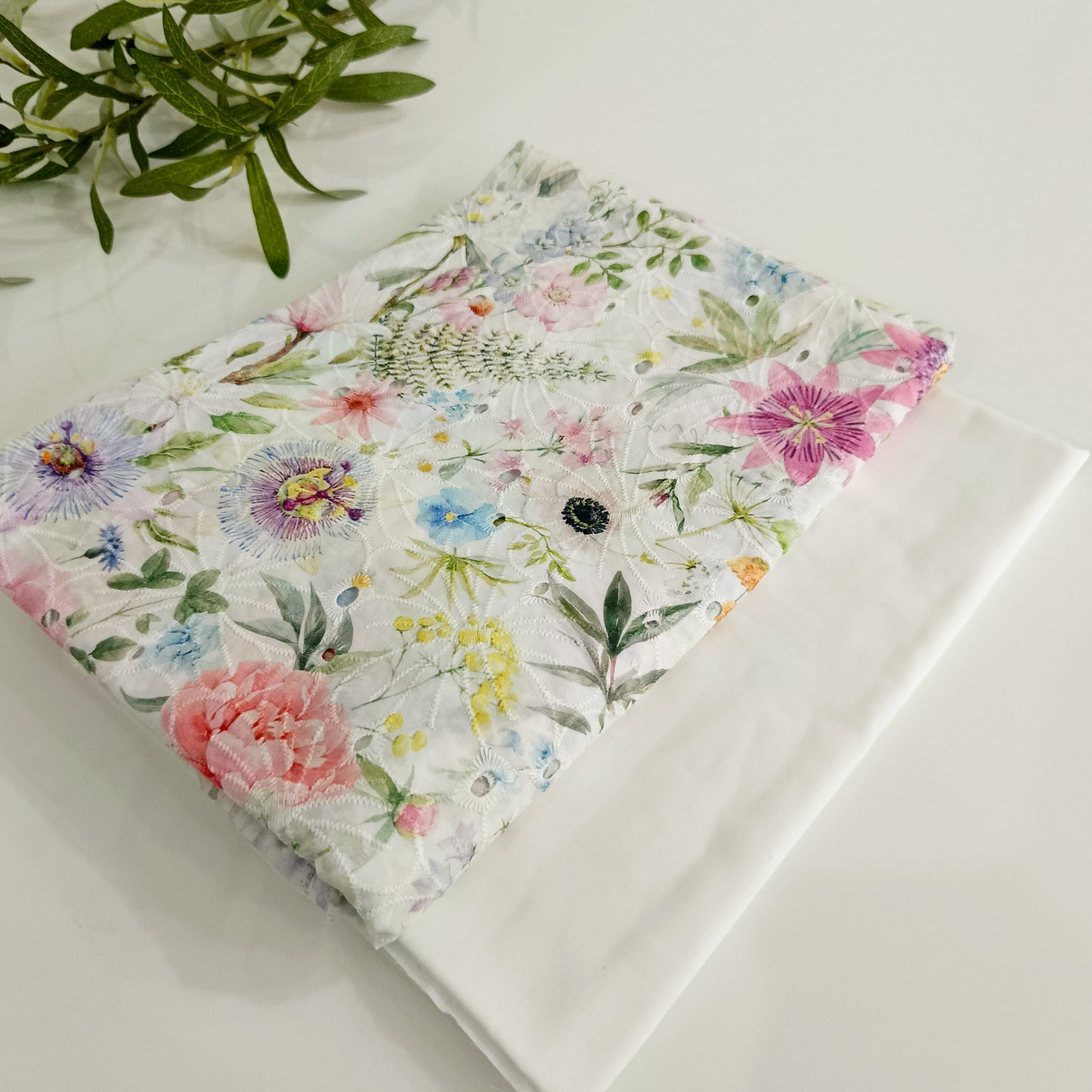 Floral-patterned fabric on a white surface with green leaves in the background
(White Multi Floral)